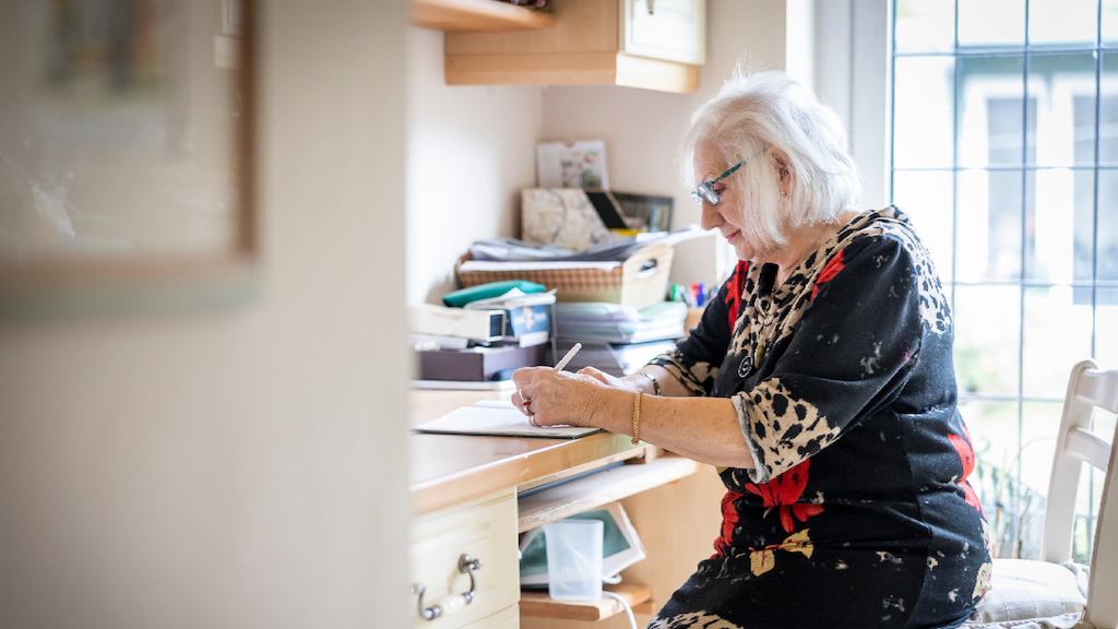 An older woman in a patterned dress writes at a desk in her home